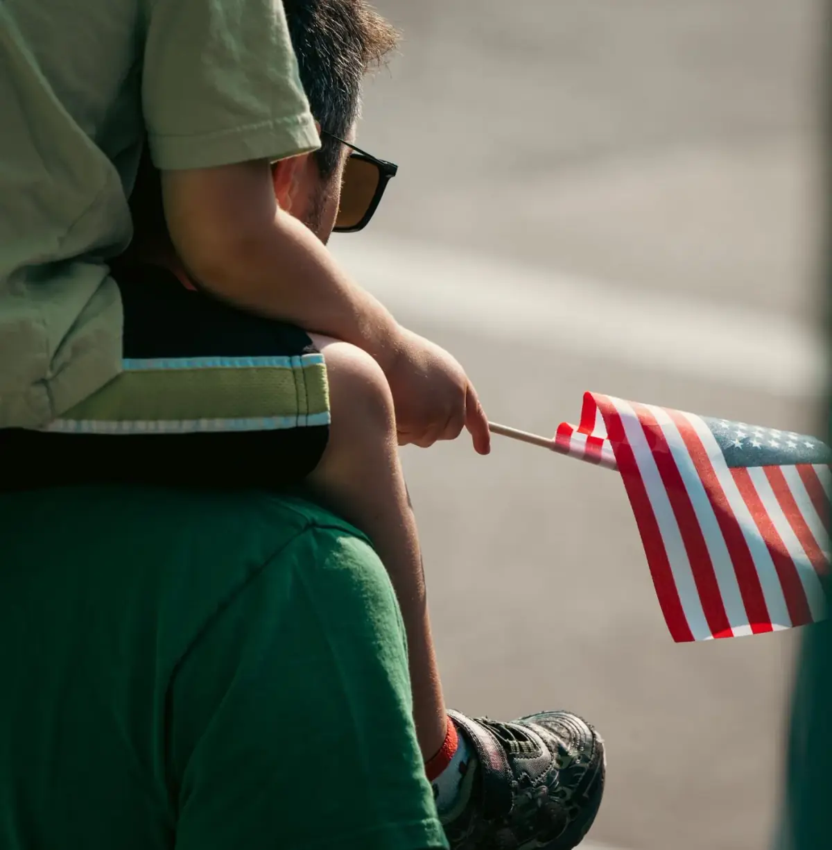 A father carries his son on his shoulders; the boy holds a United States flag
